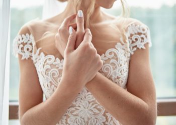 Portrait of beautiful bride in wedding dress with crossed hands standing near window in room, copy space. Bride's morning preparation